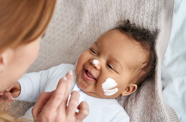 A Caucasian mother applying safe baby cream to a black baby's smiling face. Concept of safe and harmful ingredients in baby skin care products.