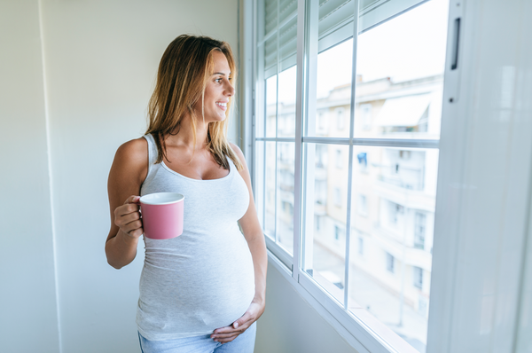 A pregnant woman holding a cup of tea and looking out a window. Concept of foods and drinks to avoid during pregnancy.