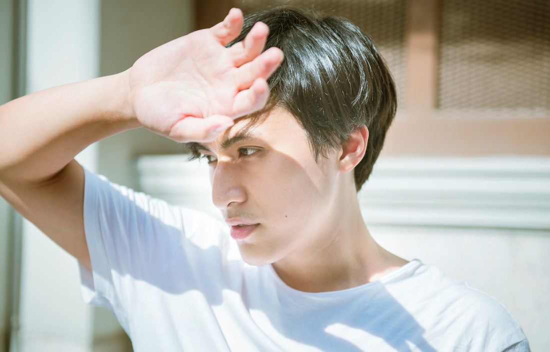 A young Asian man hides his face from the sun during a heat wave. White t shirt, outdoor space.