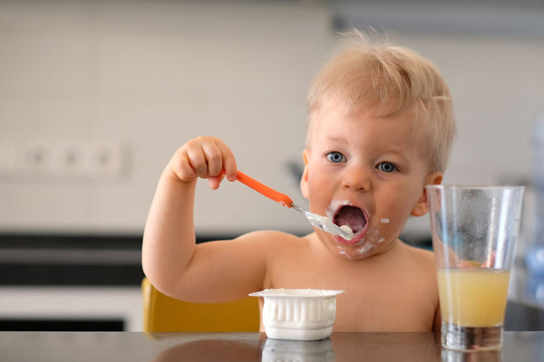 Adorable blond toddler eating yogurt with a spoon while sitting at the kitchen counter. Concept of fun and healthy summer snacks for toddlers.