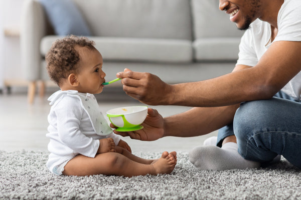 A father feeding his baby healthy food out of a bowl while sitting on the floor.