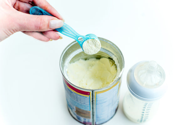 An open can of baby formula with a spoon and a baby feeding bottle nearby. 