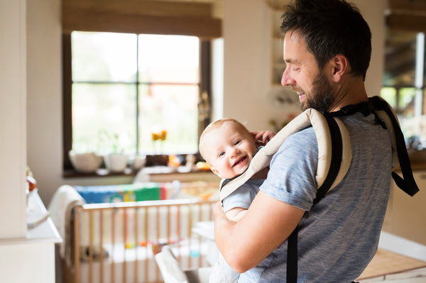 A first-time parent holding baby in a baby wrap or sling. A father with his baby, crib visible in the background.
